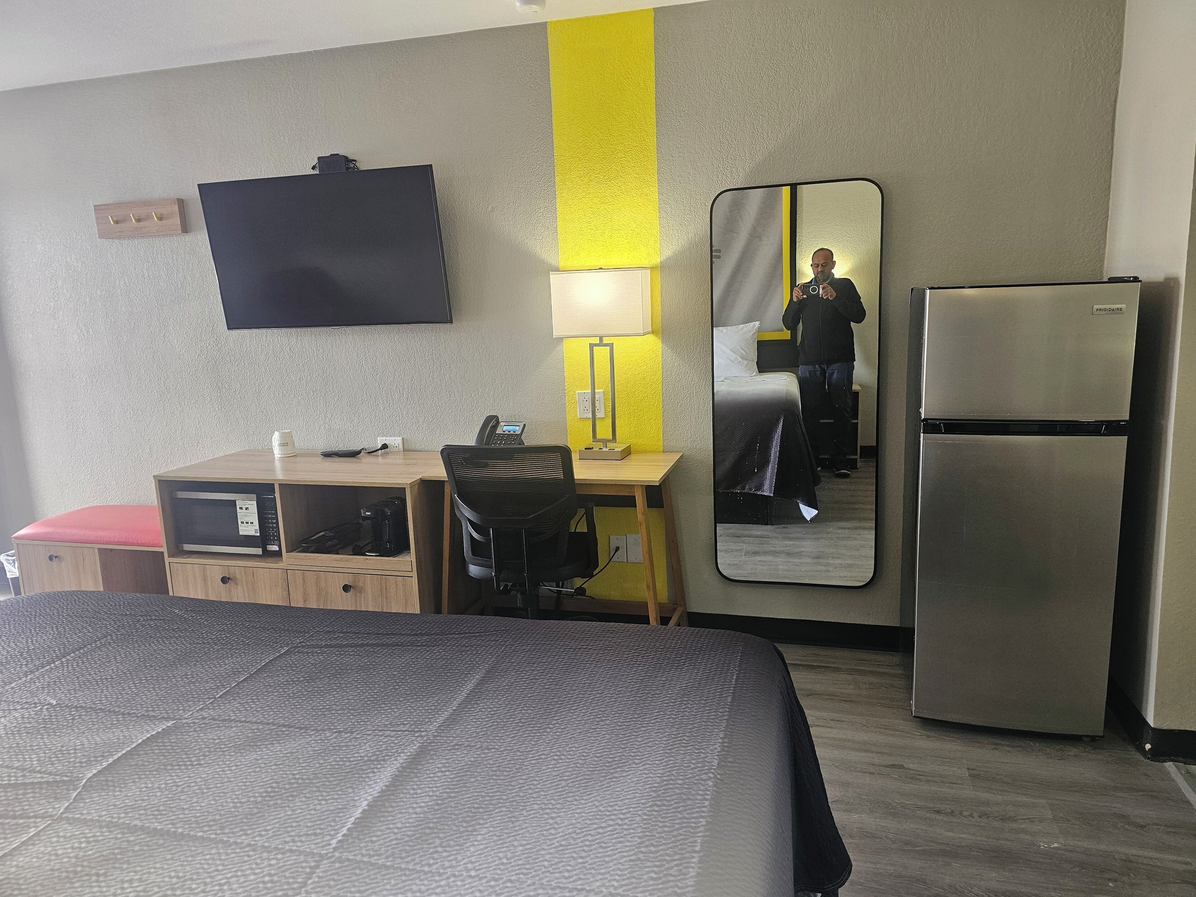 Habitación de hotel con cama, escritorio, silla, televisor de pantalla plana, refrigerador y espejo.