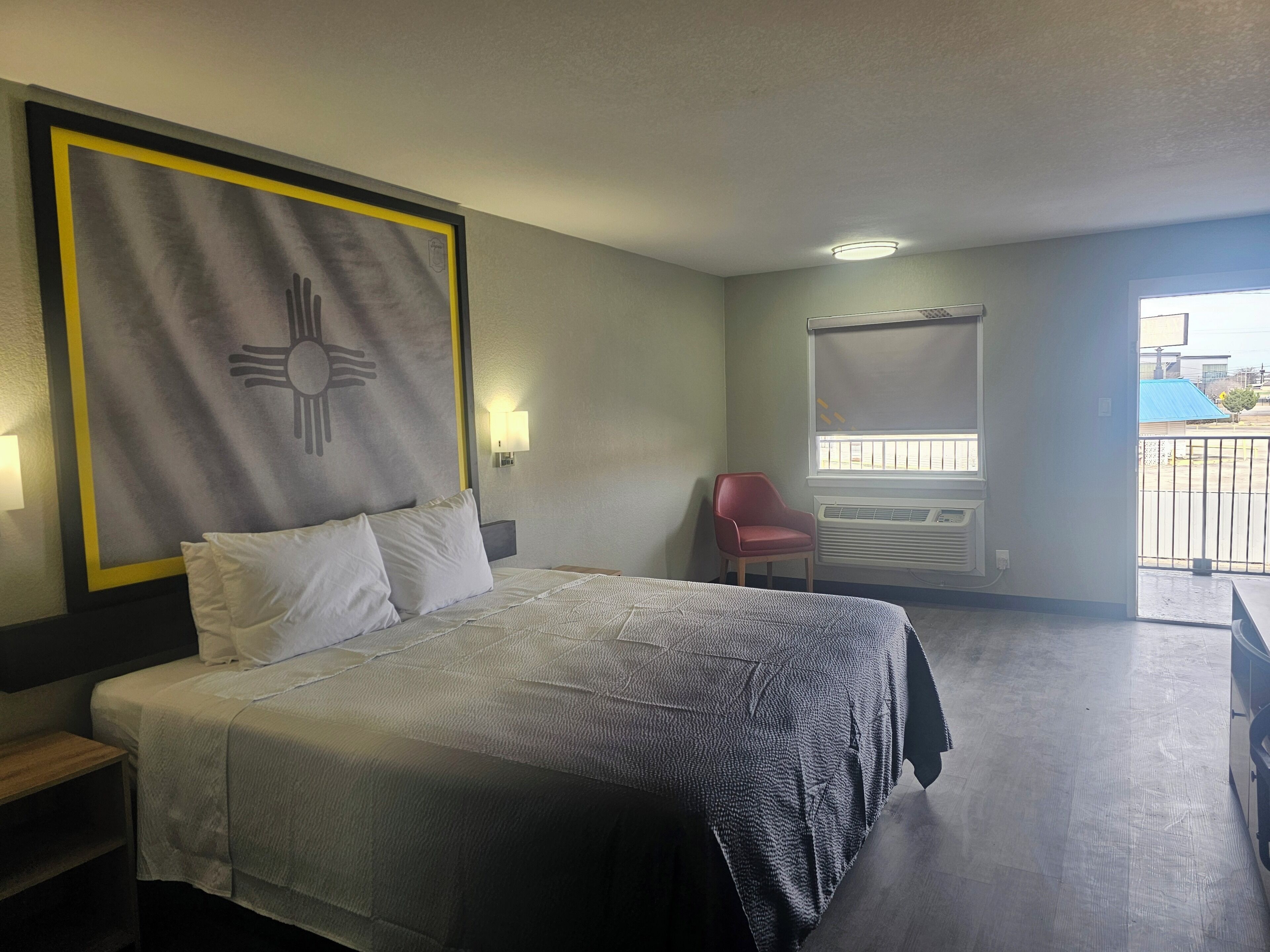 A hotel room with a bed, a red chair, a window with blinds, and a wall-mounted air conditioning unit.