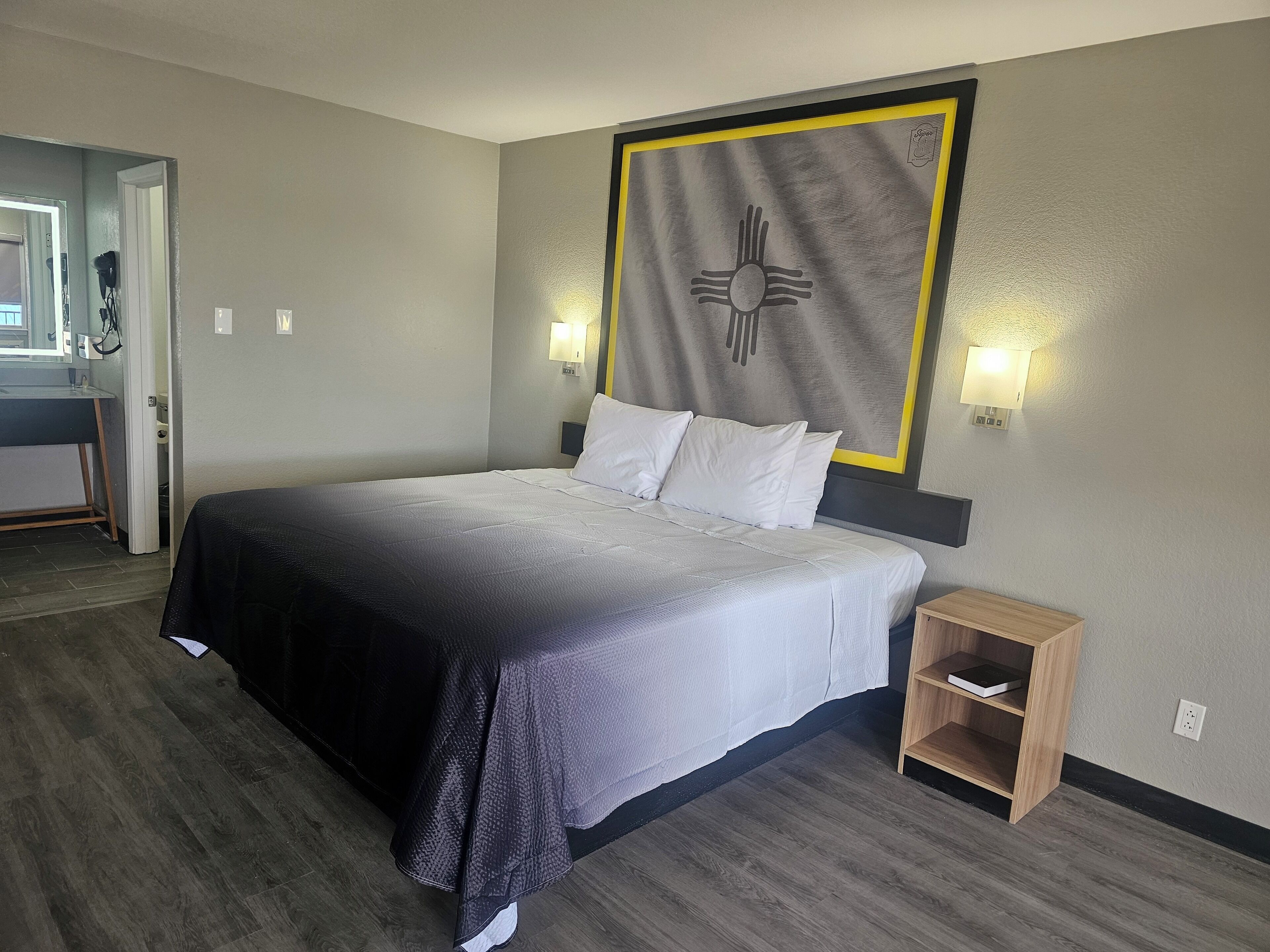 A hotel room with a bed, bedside tables, a wall-mounted artwork, and a bathroom visible through an open door.