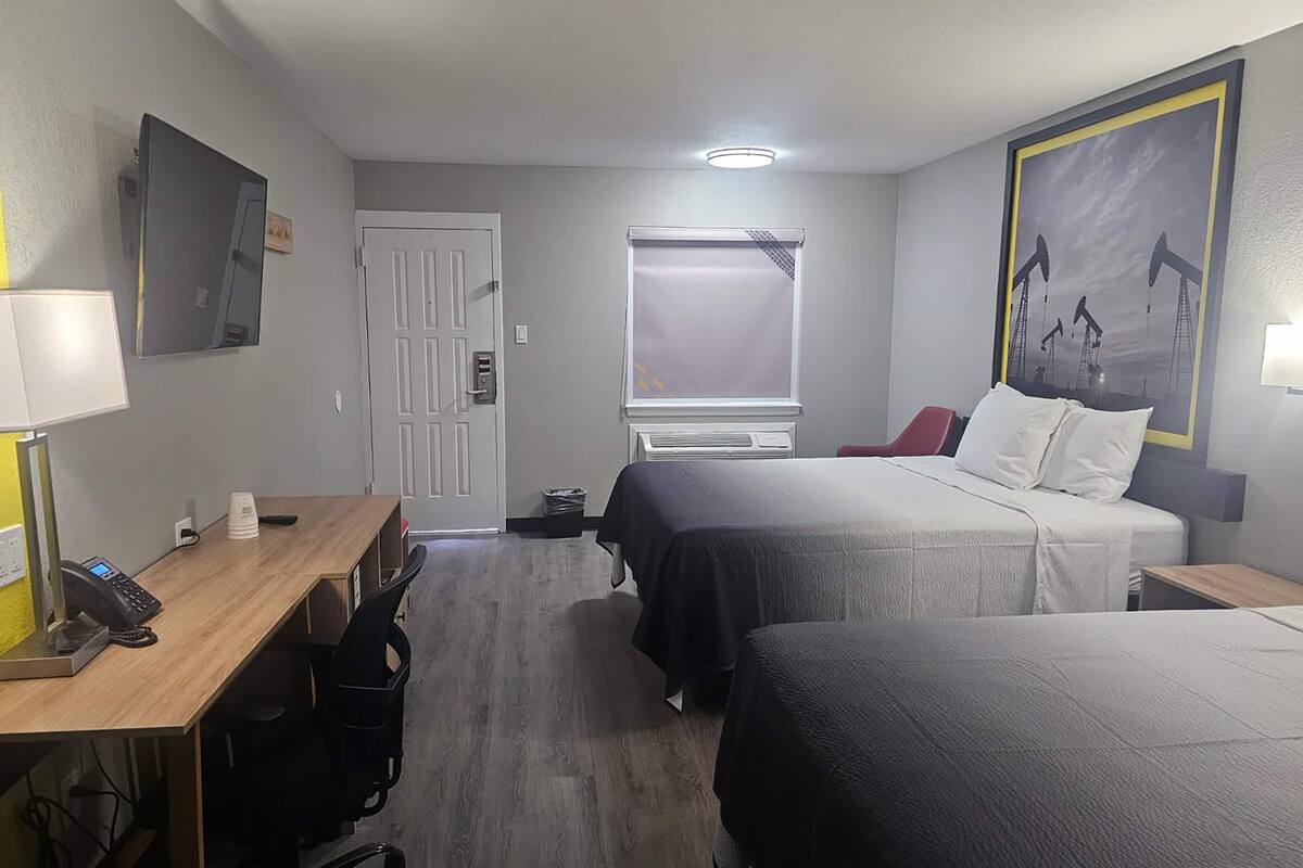 Een hotelkamer met een bureau, twee bedden, een raam met jaloezieën, een deur en een schilderij aan de muur.