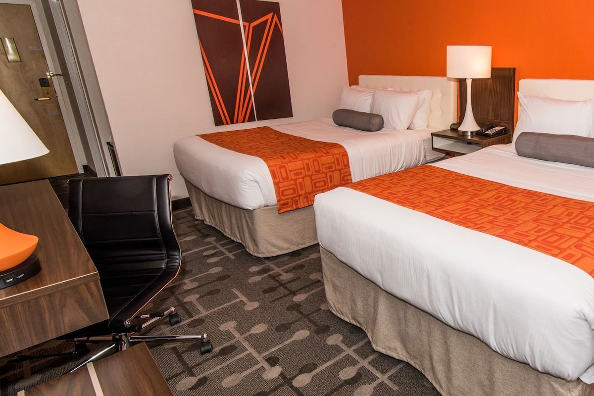 Una habitación de hotel con dos camas, un escritorio, una silla, una lámpara y una lámpara de pared.