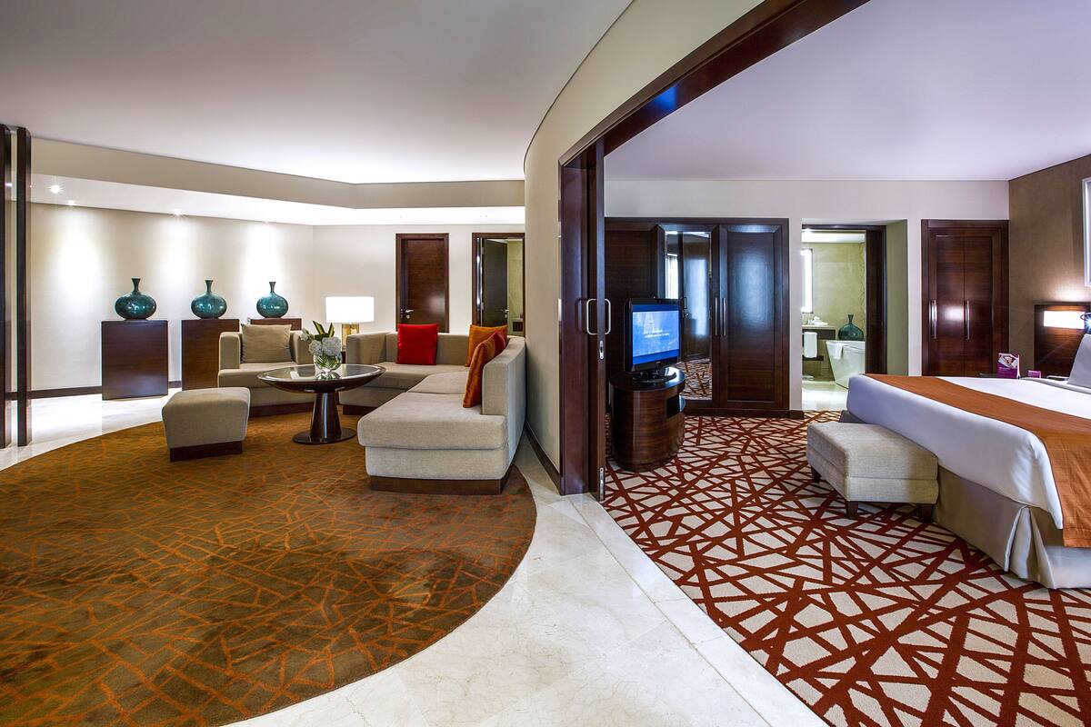 Une chambre d’hôtel avec un grand lit, une télévision, un coin salon avec une table et des chaises, et une moquette à motifs.