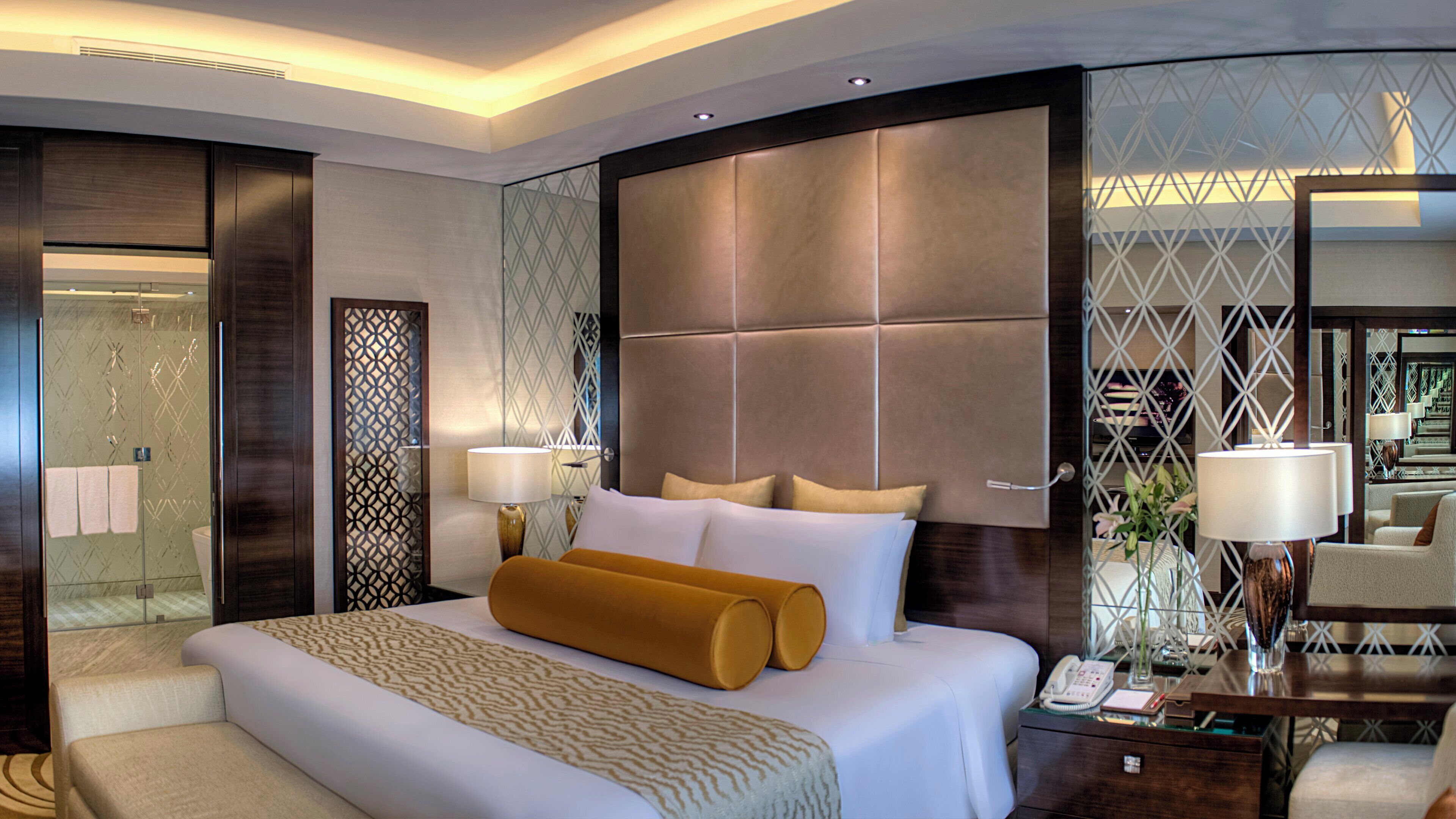 Quarto de hotel moderno com uma cama grande, uma mesa de cabeceira com abajur e um painel de vidro com padrão geométrico.