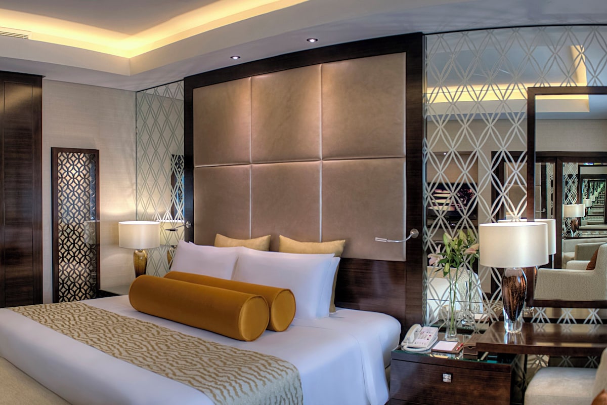 Ein Hotelzimmer mit einem großen Bett, einem Nachttisch mit Lampe, einer Glaseinteilung und Blick auf die Hotelhalle.