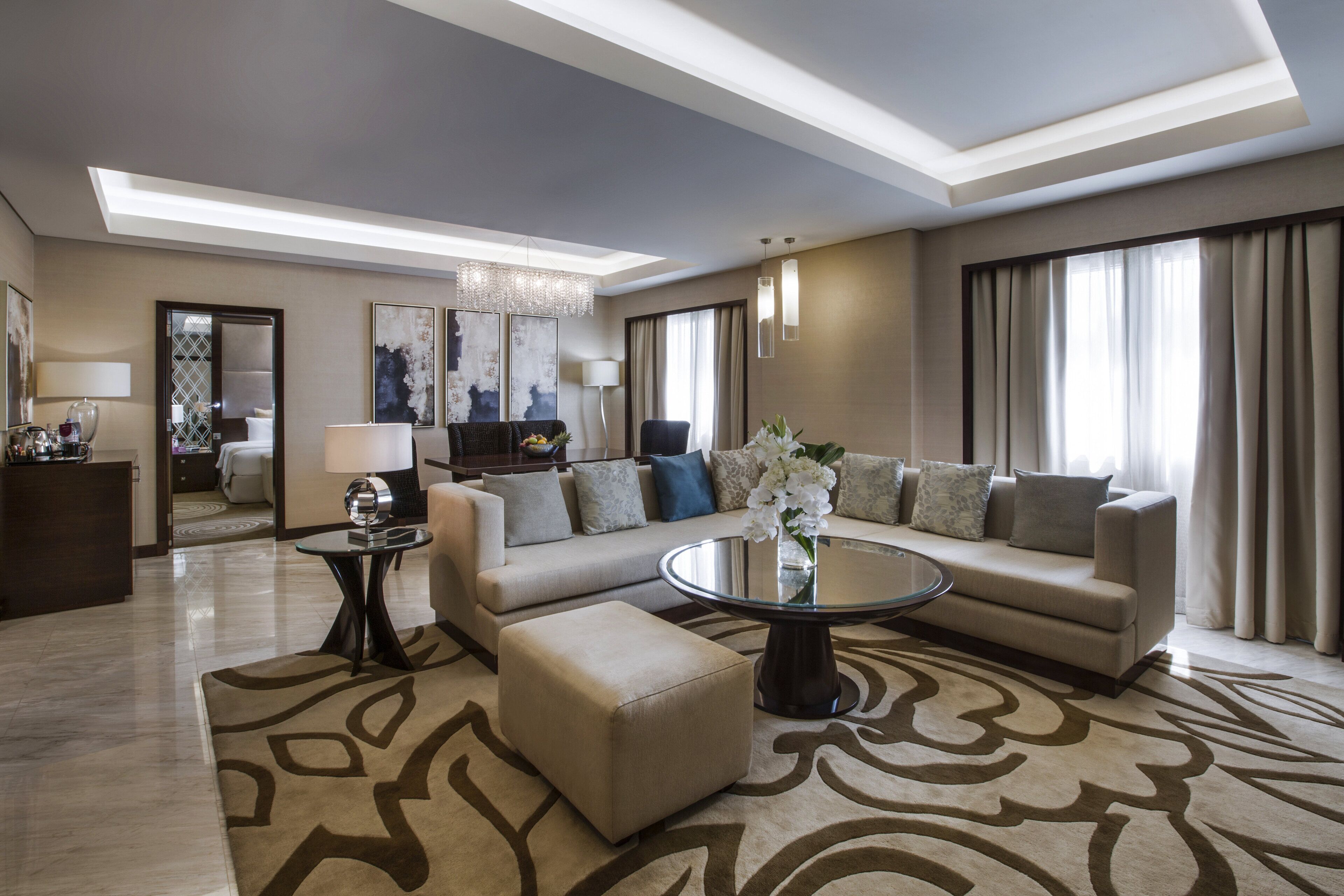 Ein modernes Wohnzimmer mit Sofa, Hocker und Couchtisch, einer Chandelierleuchtre und Blick auf das angrenzende Schlafzimmer.