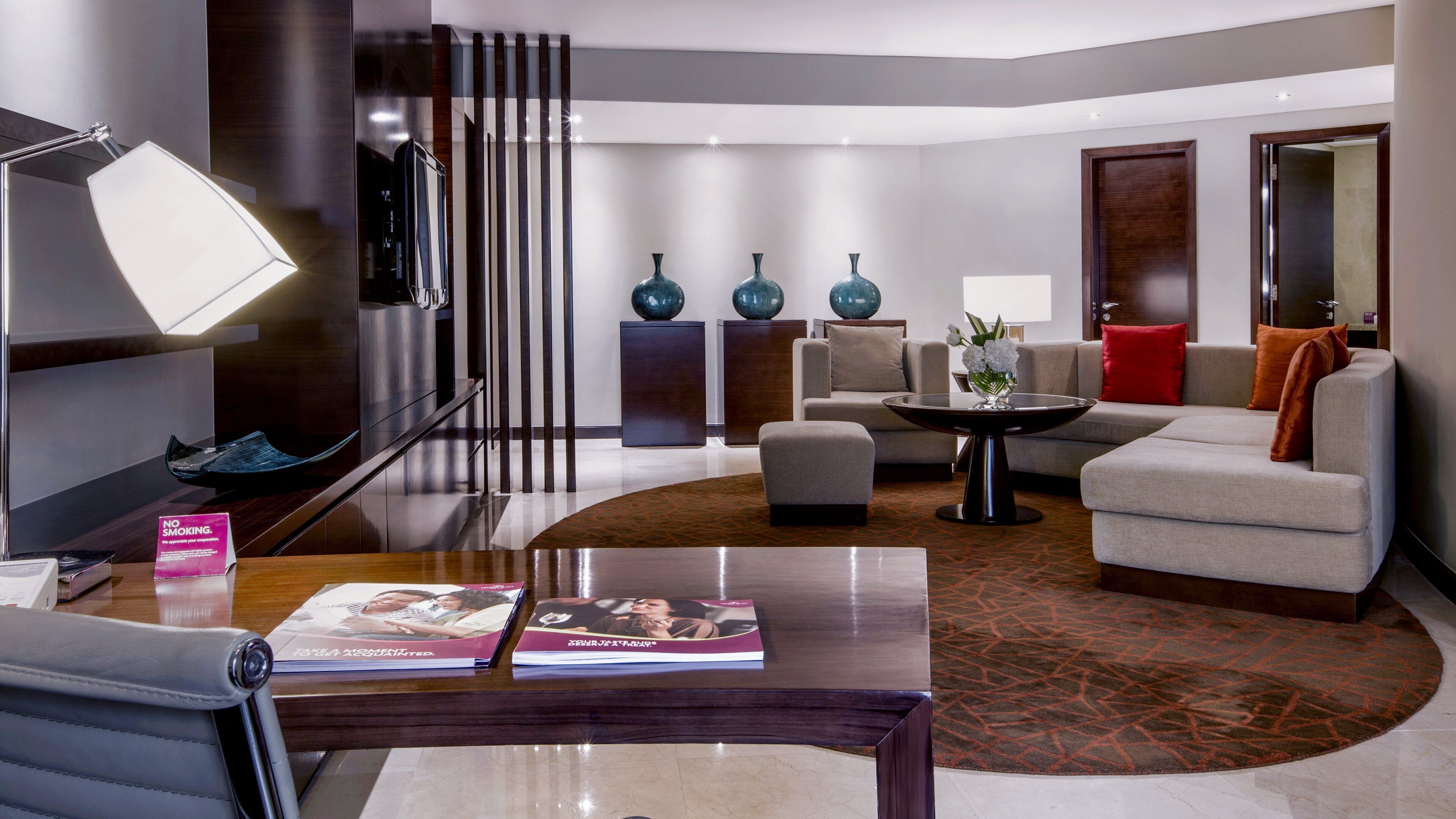 A modern living room with a sofa, armchairs, a coffee table, and a TV. There are decorative vases on a console table and a 'No Smoking' sign on the desk.