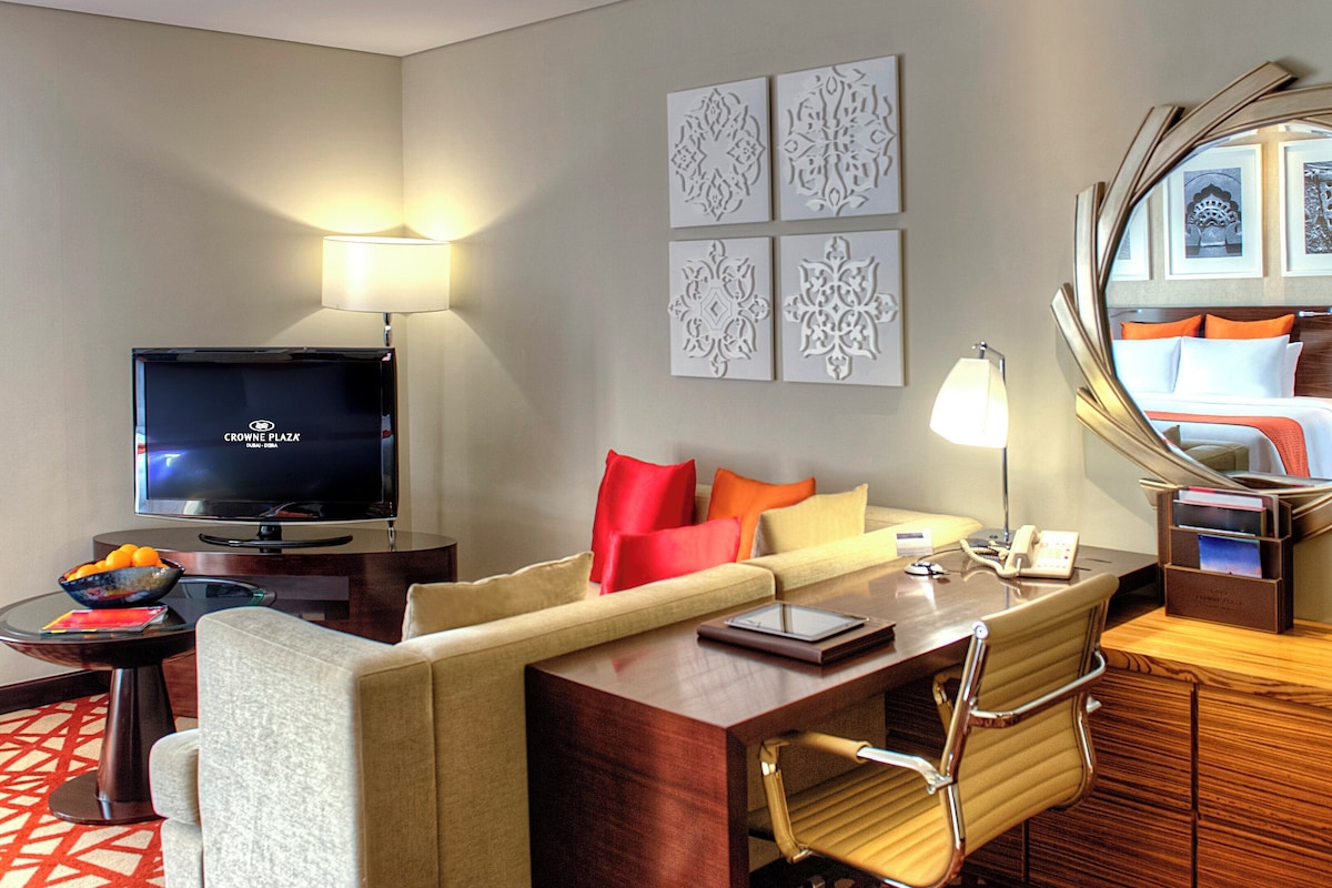 Ein Hotelzimmer mit Fernseher, einer Couch, einem Schreibtisch, einem Sessel, einem Spiegel und Bildern an den Wänden.