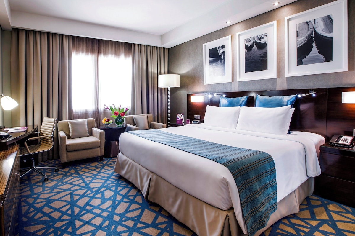 Ein Hotelzimmer mit einem großen Bett, einem Schreibtisch, einem Sessel, einer Schreibtischlampe, einem Nachttisch und einem an der Wand montierten Fernseher.
