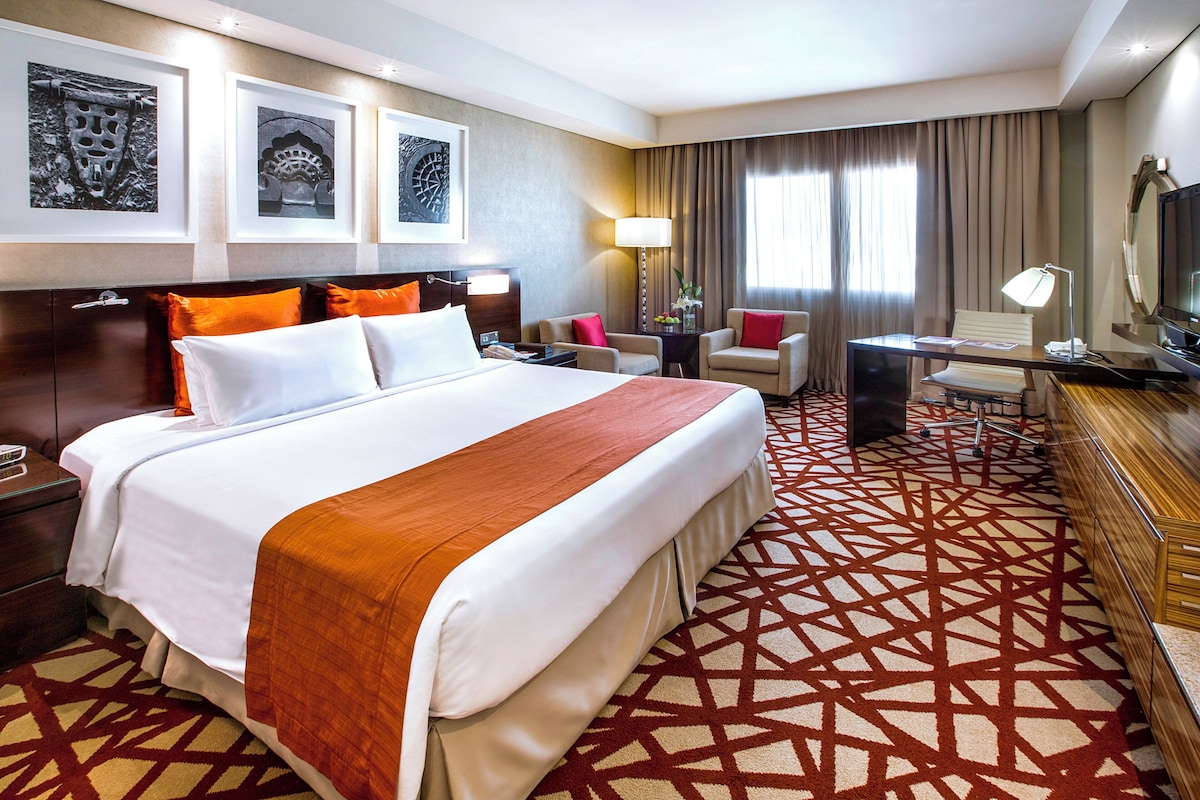 Ein Hotelzimmer mit einem großen Bett, einem Schreibtisch, einem Sessel, einem Fernseher und Bildern an der Wand.