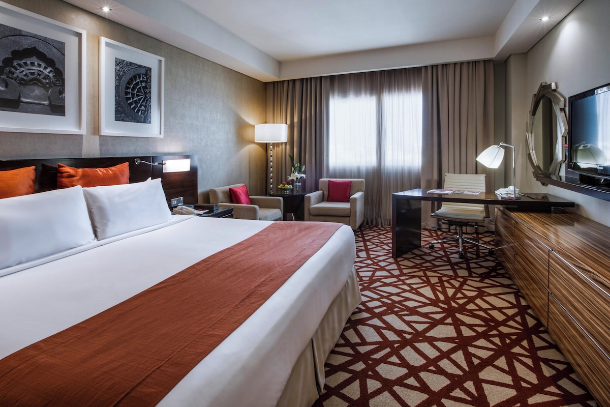 Ein Hotelzimmer mit einem großen Bett, einem Schreibtisch, einem Sessel, einem Fernseher und einem gemusterten Teppich.