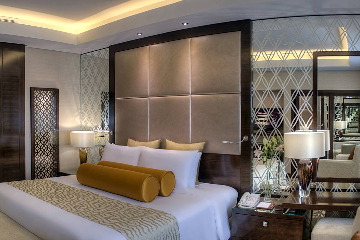 Une chambre d’hôtel moderne dotée d’un grand lit, d’une table de chevet avec une lampe et d’une cloison en verre ornée d’un motif géométrique.