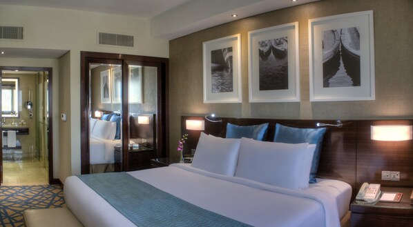 Une chambre d’hôtel avec un grand lit, un bureau avec un téléphone et un miroir. Trois tableaux encadrés sont accrochés au mur, au-dessus du lit.