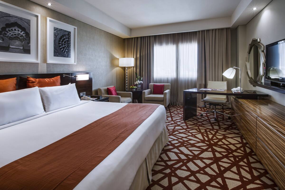 Une chambre d’hôtel avec un grand lit, deux fauteuils, un bureau, une télévision et une moquette à motifs.