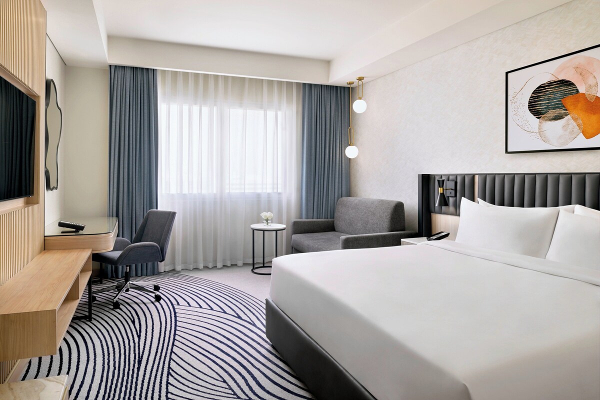 Ein modernes Hotelzimmer mit einem großen Bett, einem Schreibtisch mit Stuhl, einem kleinen Beistelltisch und einem Fernseher.