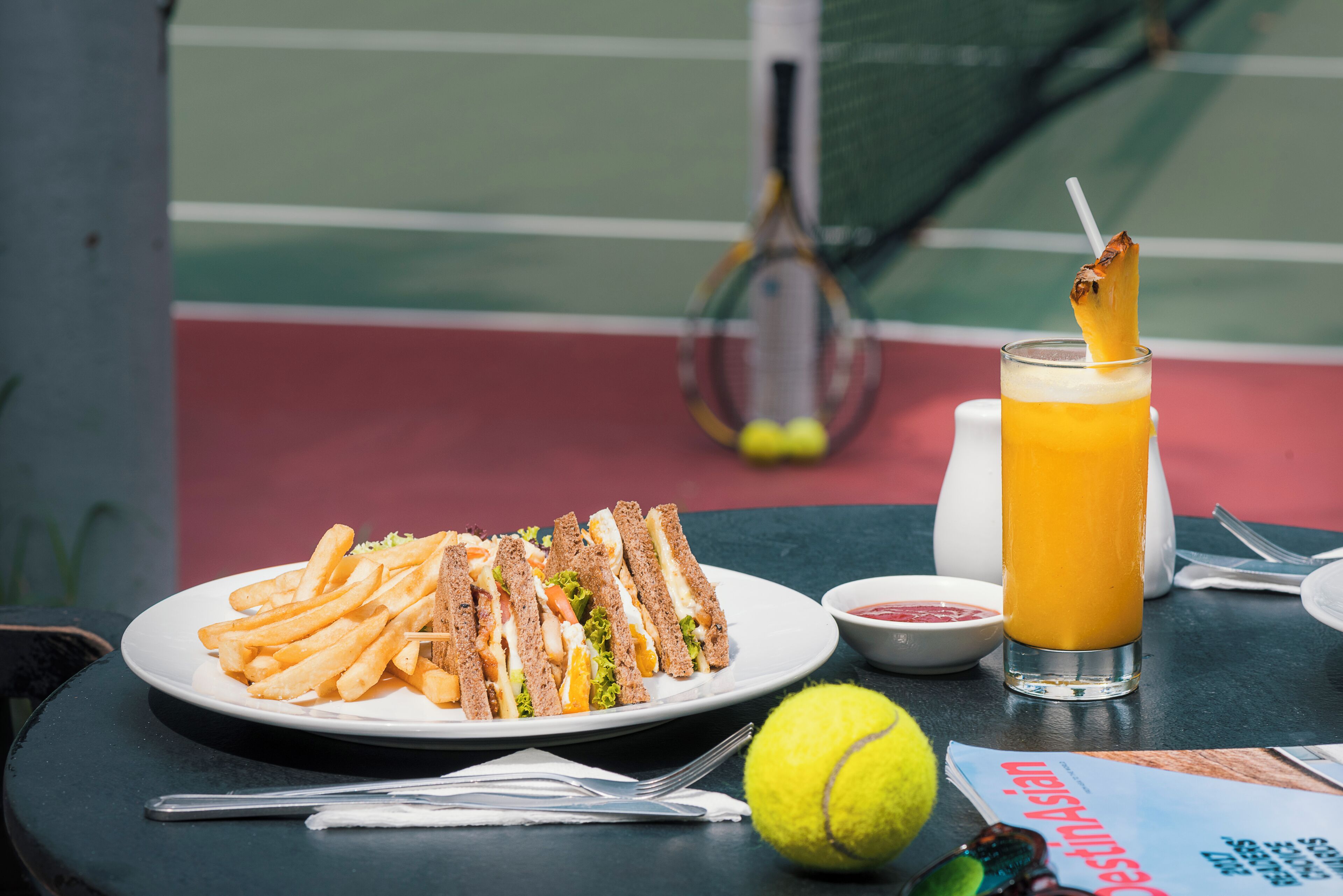 Una cancha de tenis con una mesa dispuesta para comer, que incluye un sándwich, papas fritas, un vaso de jugo y una pelota de tenis.