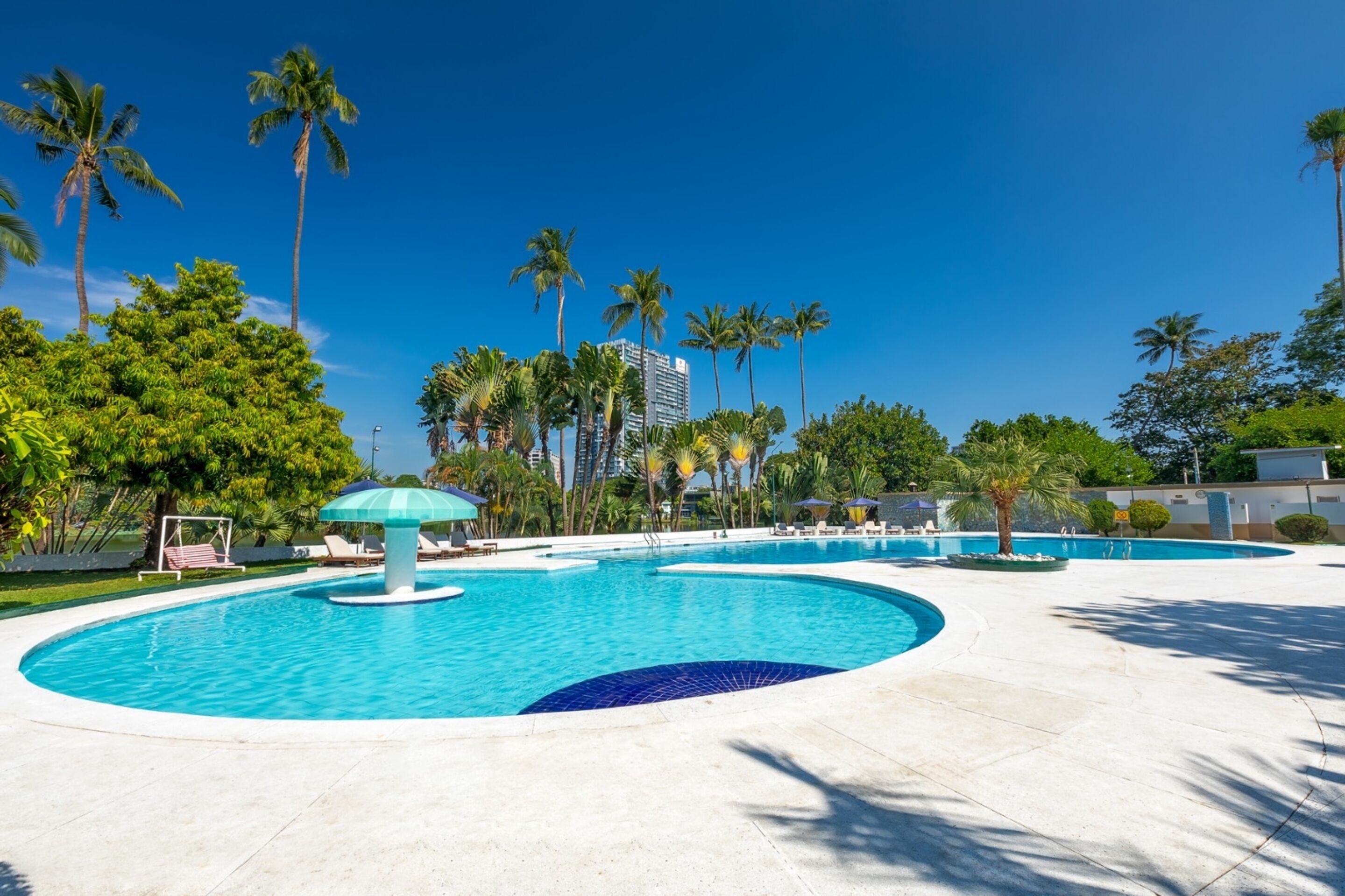 Una amplia piscina al aire libre con palmeras y sillas de descanso, rodeada de un césped bien cuidado y un edificio al fondo.