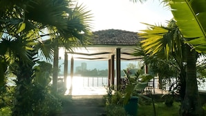 A covered outdoor area with a view of a body of water, surrounded by lush greenery and a tiled roof structure.