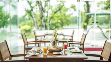 Una mesa de comedor dispuesta con alimentos para el desayuno, como pan, fruta y una jarra, en una habitación con grandes ventanales y sillas de madera.