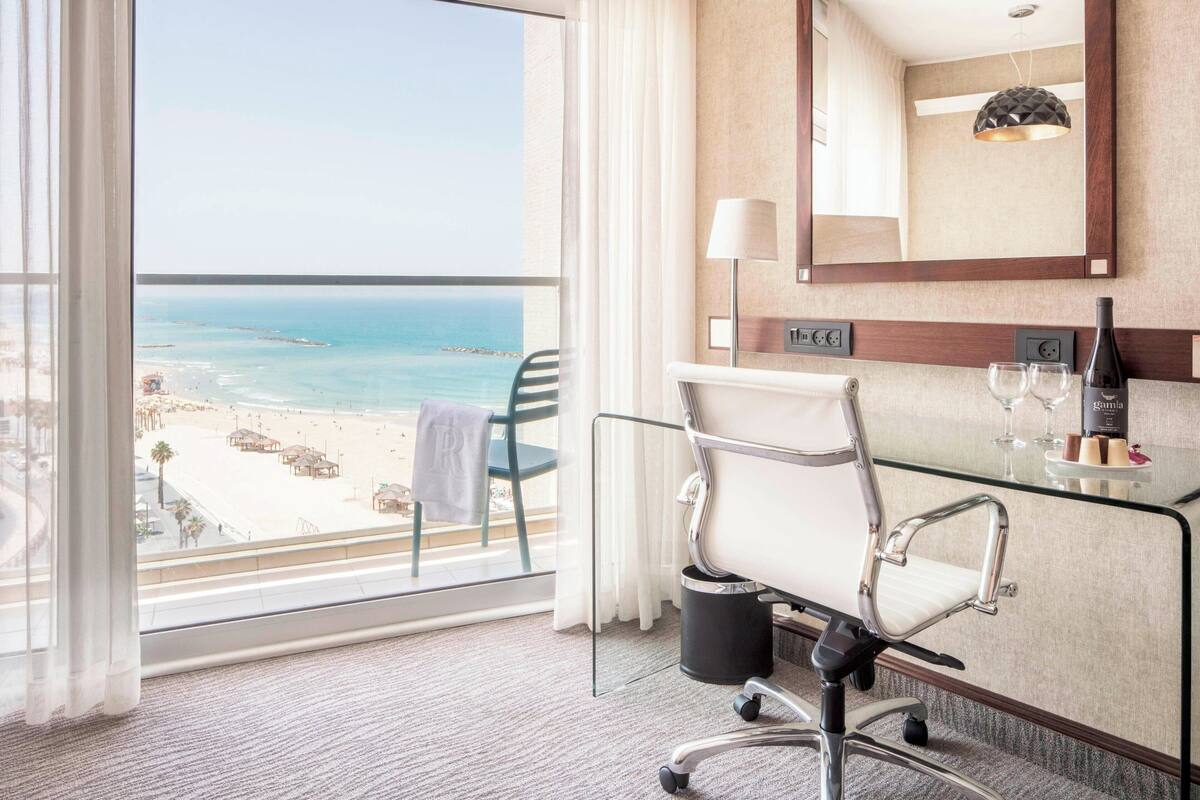 A hotel room with a view of the beach, a glass desk with a chair, a lamp, and a minibar.