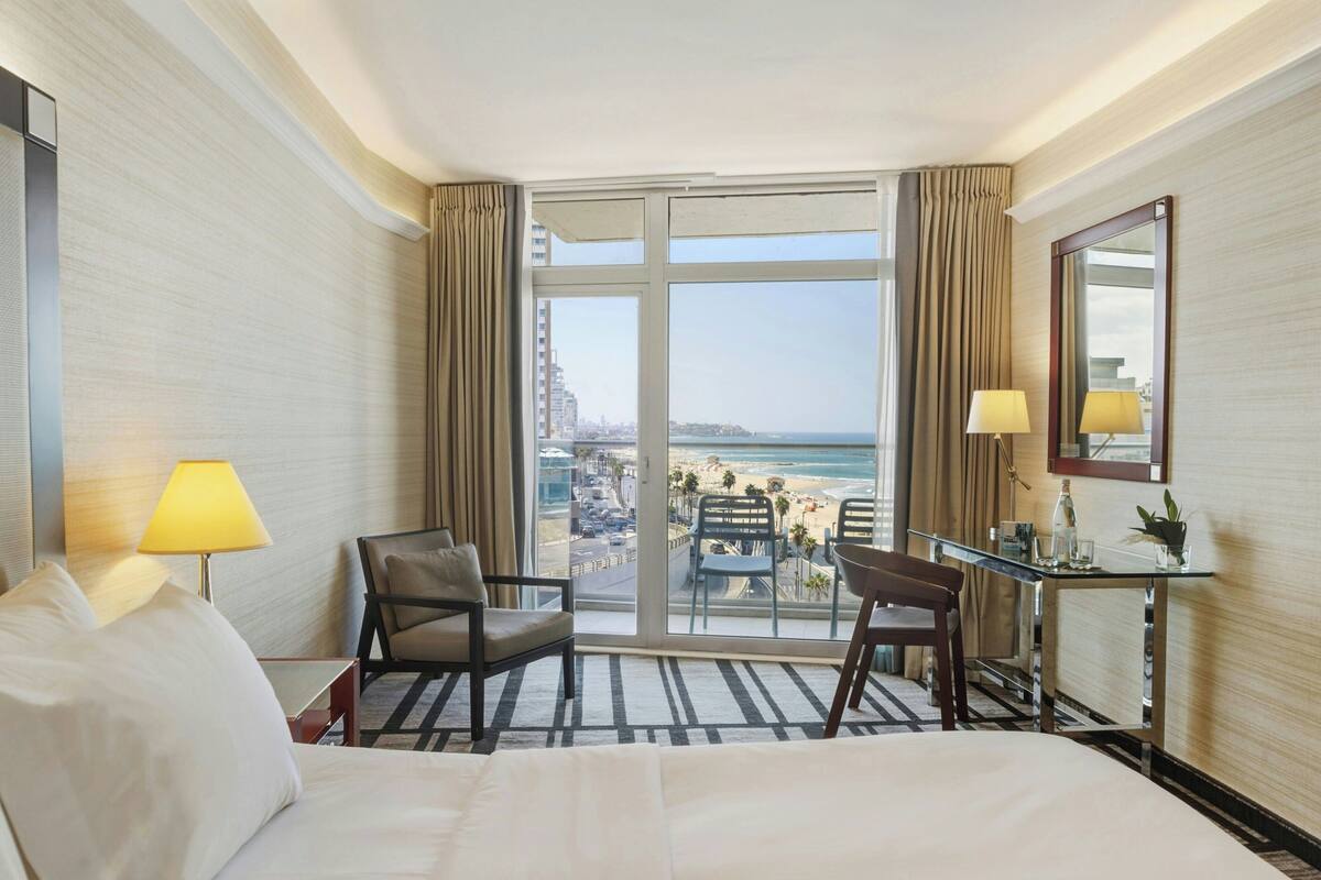 A hotel room with a large window, a bed, two chairs, a desk, and a mirror.