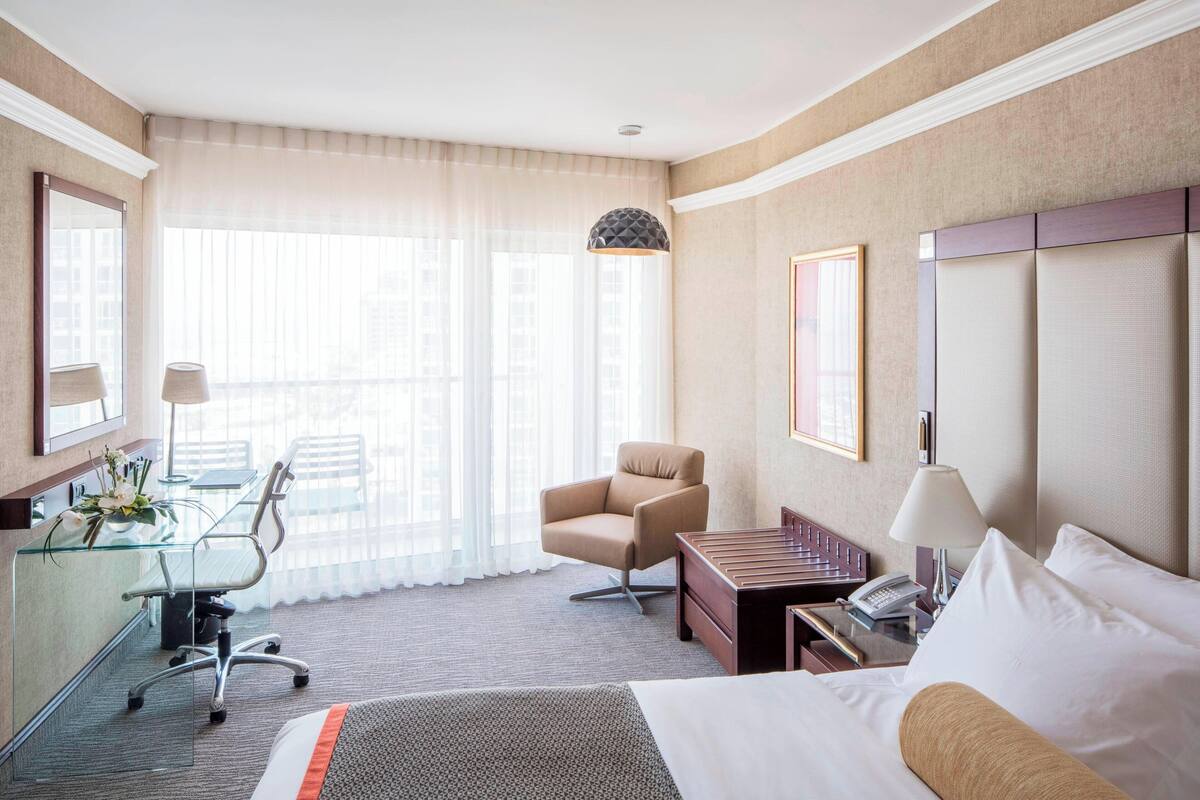 A hotel room with a bed, a desk with a chair, a sofa, a lamp, and a window with curtains.