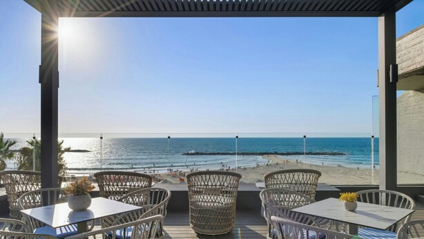 Une terrasse dotée de chaises et de tables en osier, offrant une vue sur une plage animée où l’on aperçoit des gens et des bateaux.