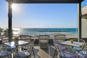 A terrace with wicker chairs and tables, overlooking a beach with people and boats.