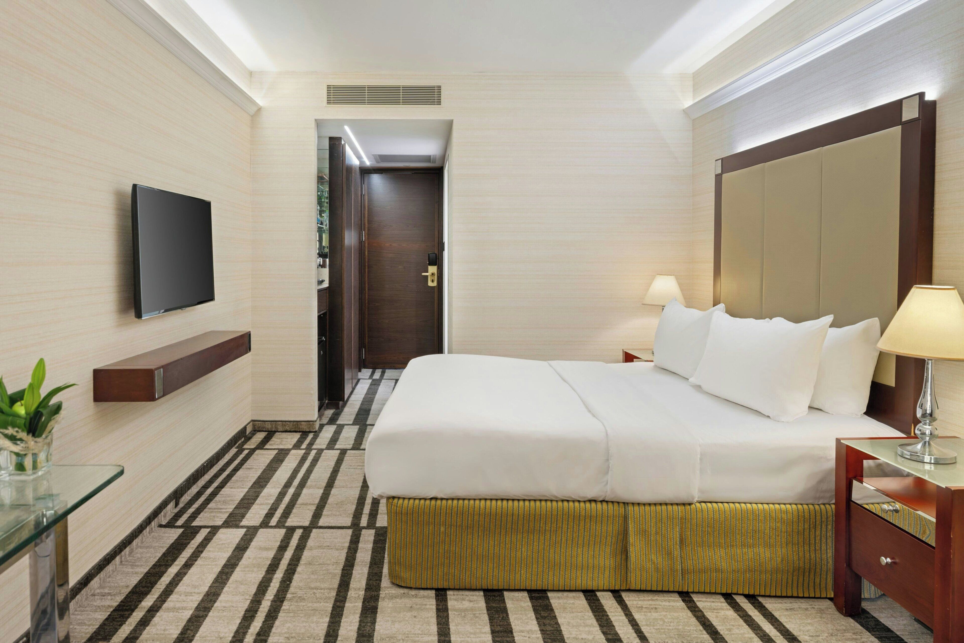 A hotel room with a bed, bedside table, lamp, TV, and a small plant.