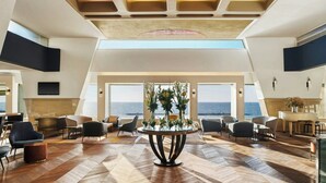 Un hall d’entrée spacieux avec un plafond haut, une table ronde centrale agrémentée de compositions florales et une vue sur la mer grâce à de grandes fenêtres.