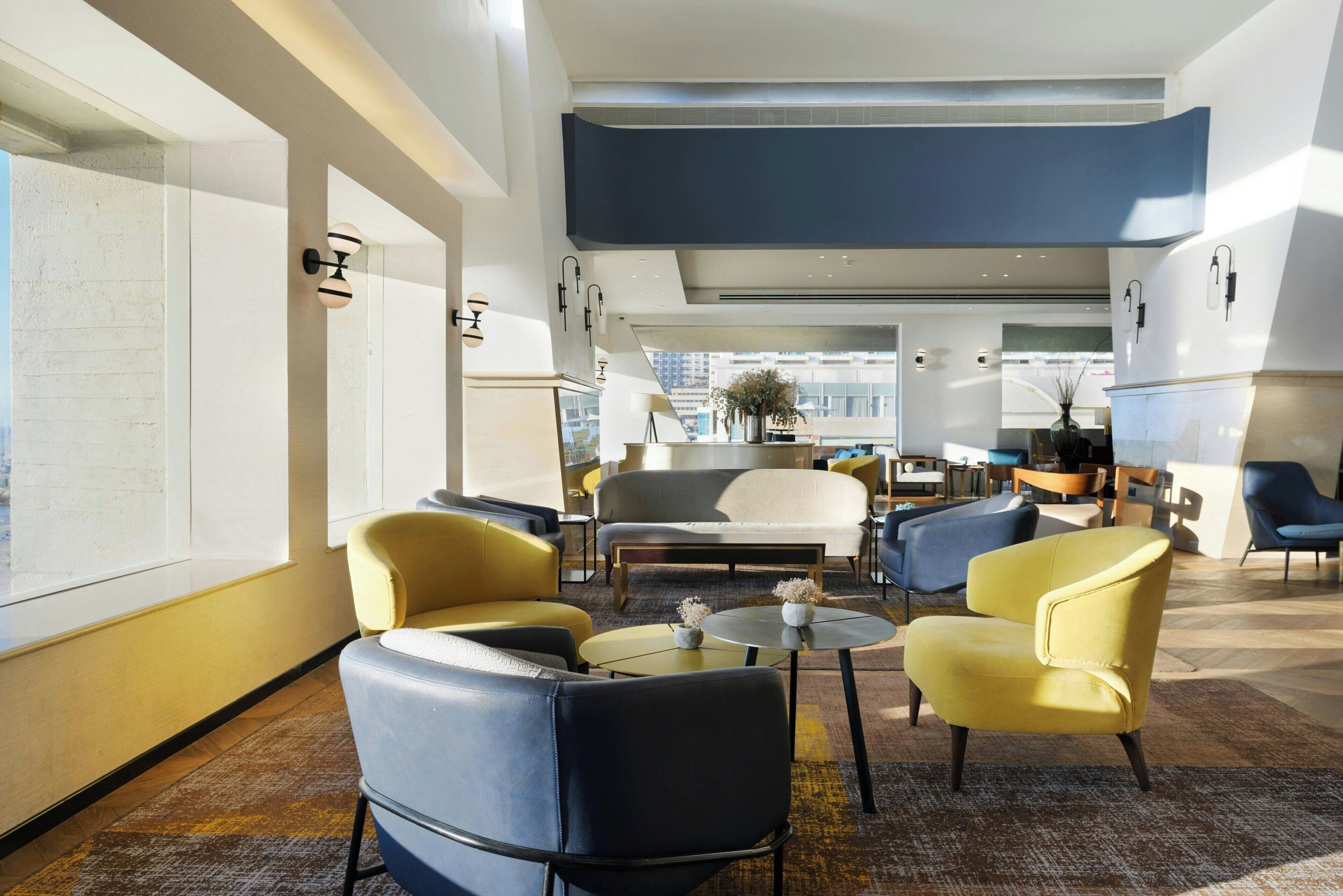 A modern hotel lobby with a high ceiling, large windows, and a mix of seating areas including sofas and armchairs.