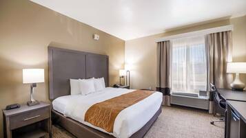 Habitación de hotel con una cama grande, un escritorio, una silla, una lámpara, un calentador y una ventana con cortinas.