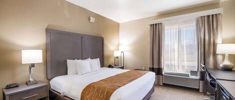 A hotel room with a large bed, a desk, a chair, a lamp, a heater, and a window with curtains.