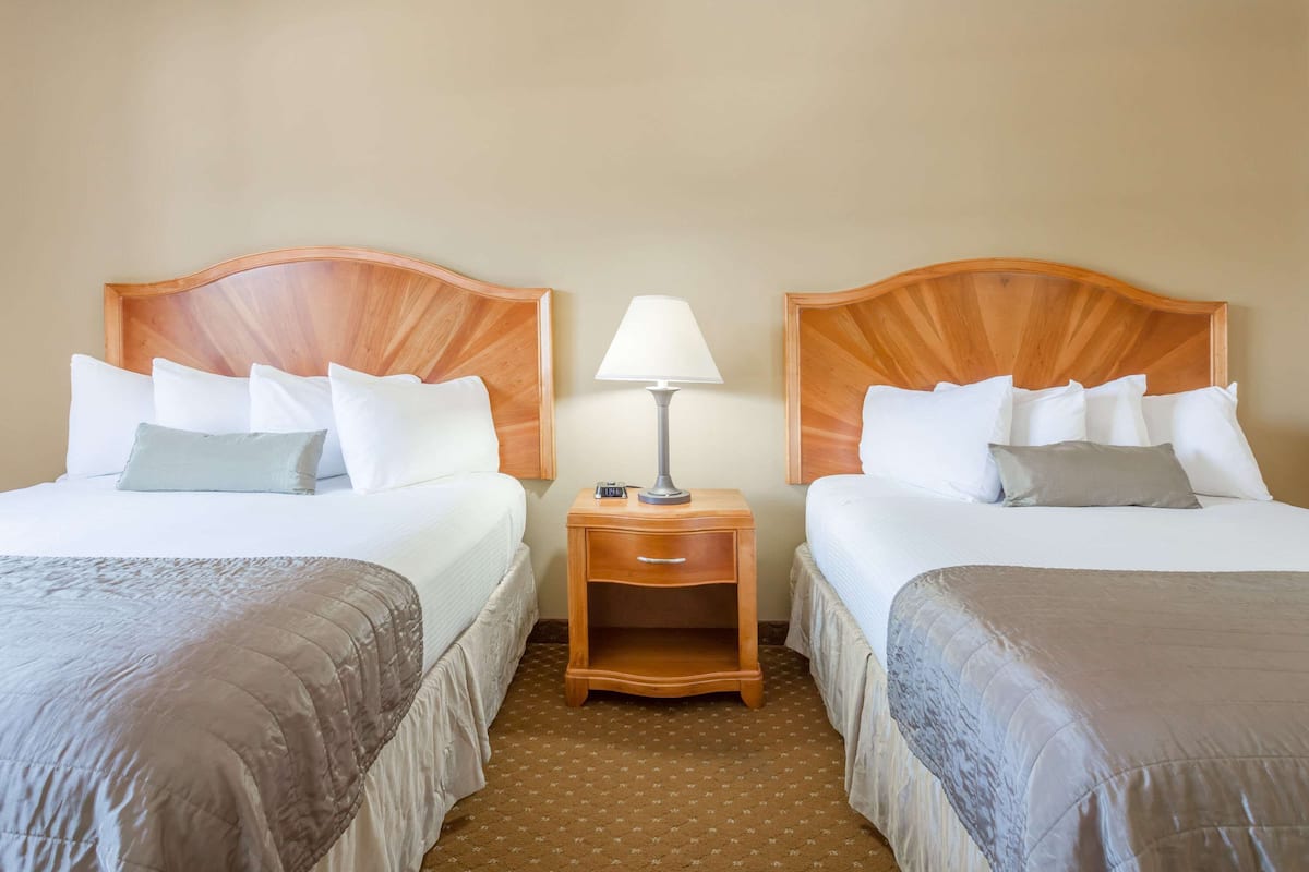 A hotel room with two beds, each with a wooden headboard, a nightstand with a lamp, and a small drawer.