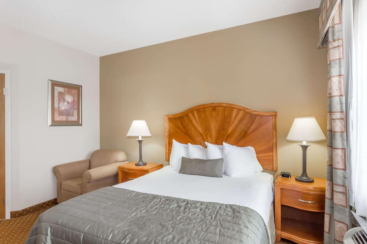 A hotel room with a large bed, two bedside tables with lamps, a chair, a framed picture on the wall, and a window with curtains.