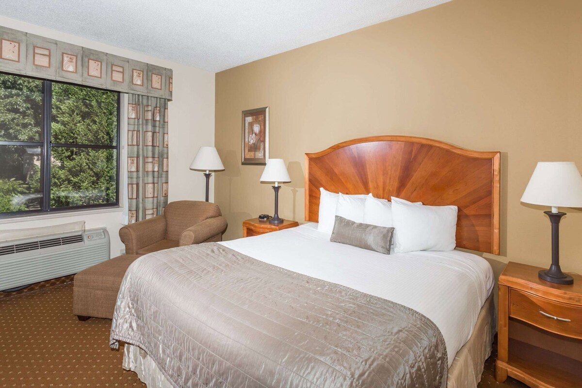 Quarto de hotel com uma cama grande, duas mesas de cabeceira com luminárias, uma poltrona, uma janela com cortinas e um radiador.