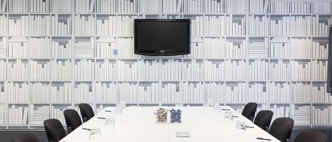 Un salón de conferencias con una mesa blanca, sillas negras, un televisor Samsung montado en la pared y una estantería al fondo.