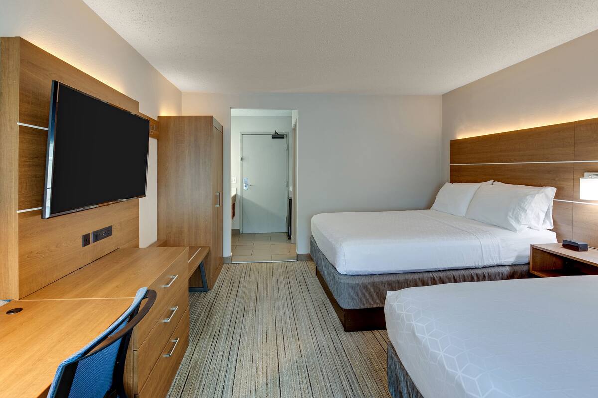 A hotel room with a wooden desk, a flat-screen TV, two beds, a walk-in closet, and a hallway leading to a bathroom.