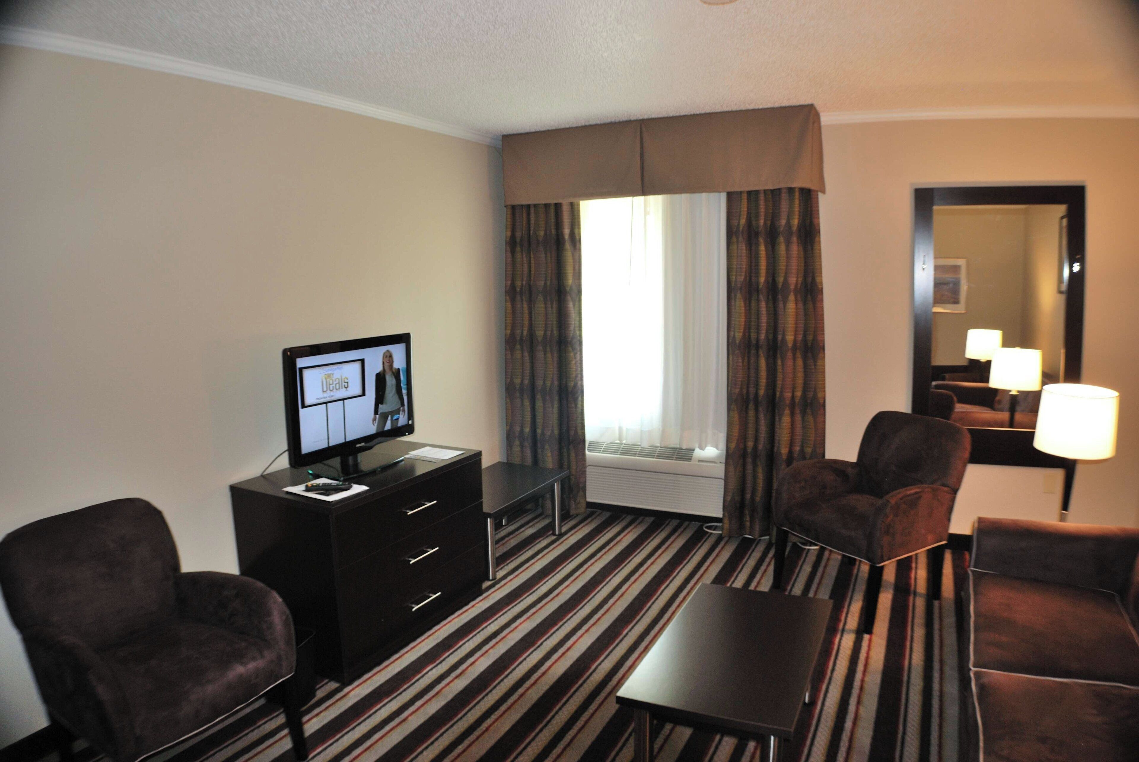 Habitación de hotel con cama, televisor, escritorio, dos sillas, cómoda y ventana con cortinas.