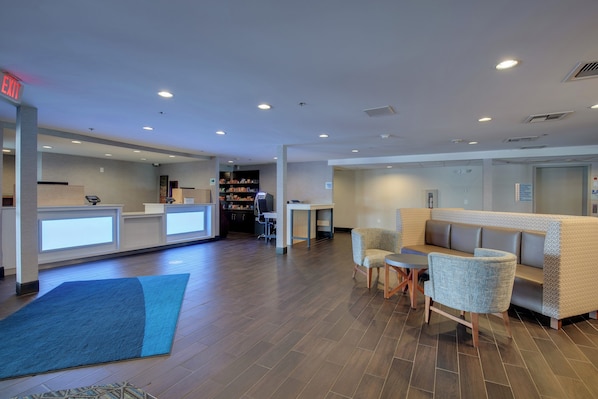 A hotel lobby with a reception desk, seating area, and a kitchenette in the background.