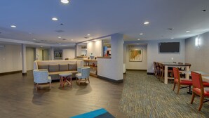 A hotel lobby with a sofa, chairs, a dining table, and a TV mounted on the wall. - Holiday Inn Express W-I40/Whitebridge Road by IHG (Nashville)
