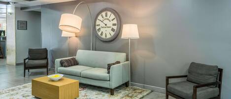 A modern lobby with a grey sofa, two armchairs, a wooden coffee table, and a large wall clock.