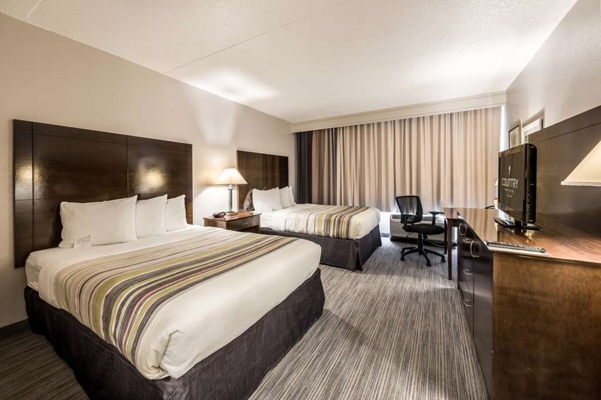 A hotel room with two beds, a desk, a chair, a TV, and a lamp.