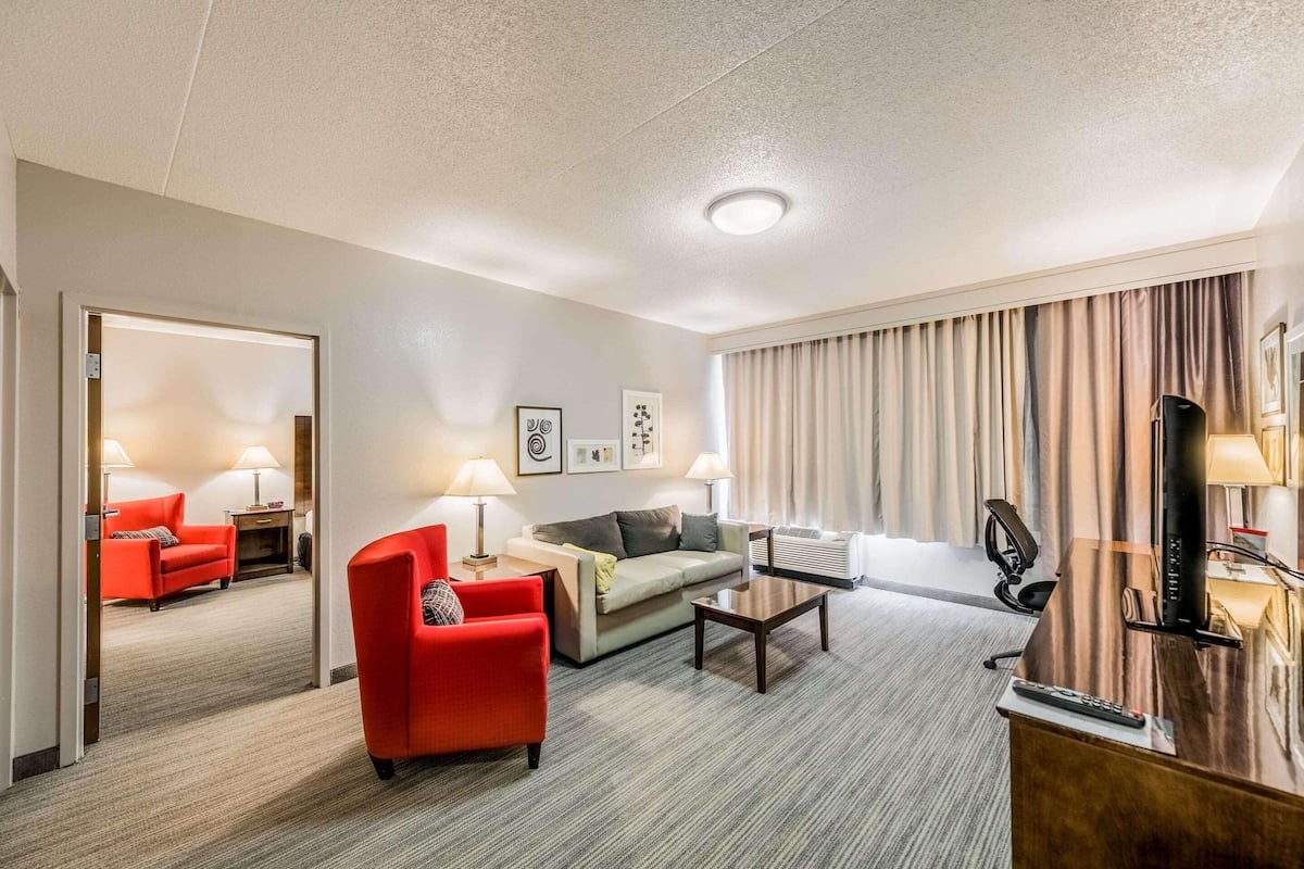 Une chambre d’hôtel avec un fauteuil rouge, un canapé, une table basse, une télévision et un bureau avec une lampe.