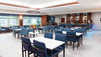 A hotel dining area with multiple tables and chairs, a reception desk, and a counter with a blue backlit display.