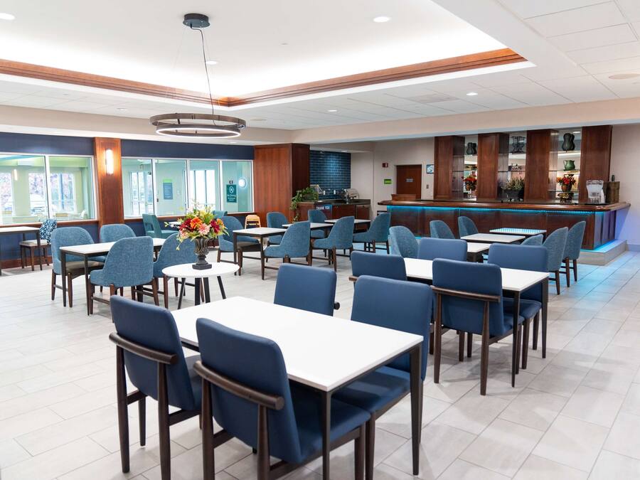 A hotel dining area with multiple tables and chairs, a reception desk, and a counter with a blue backlit display.