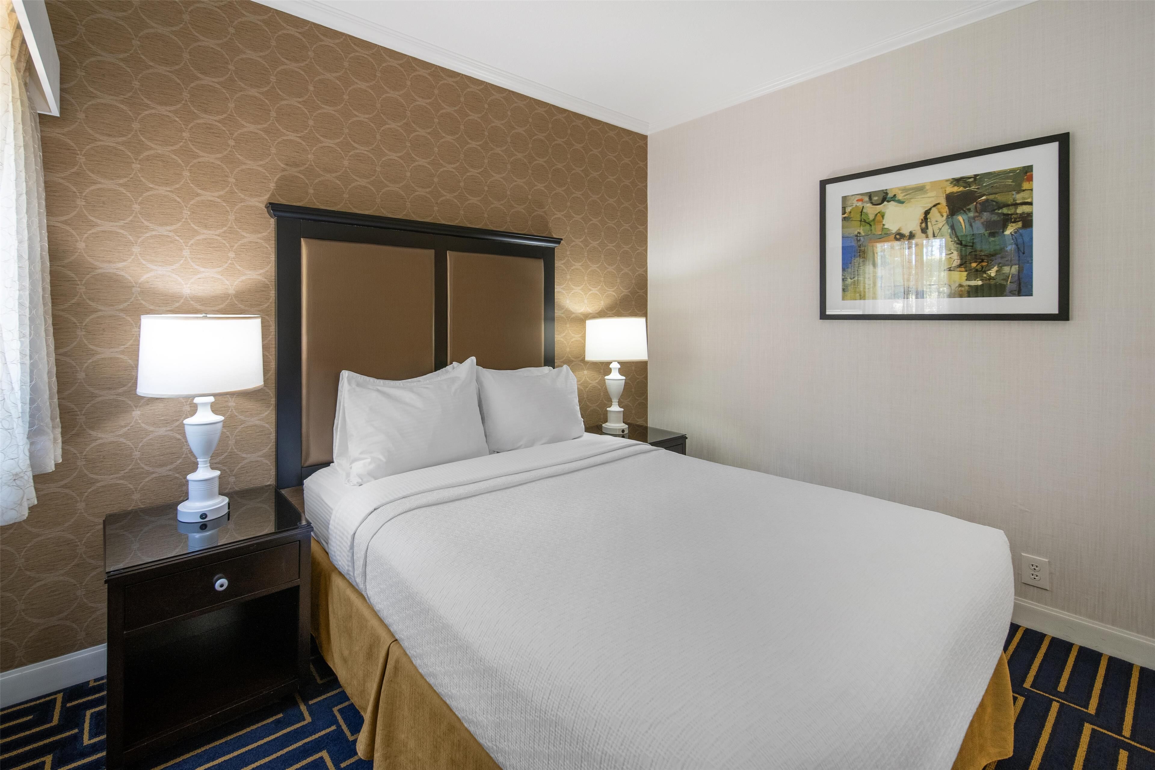 A hotel room with a large bed, two bedside tables with lamps, a patterned wallpaper, and a framed artwork on the wall.