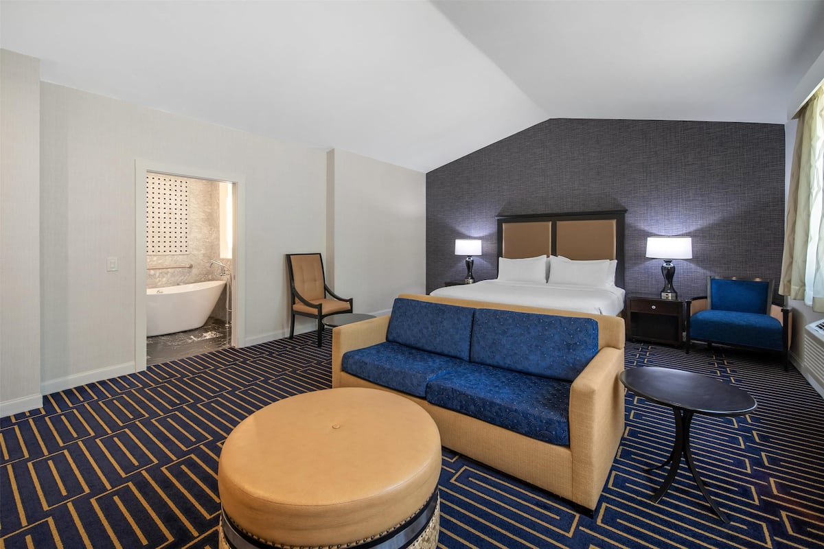 A hotel room with a bed, a sofa, a small table, a chair, and a bathroom with a bathtub.