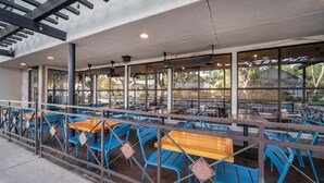 Ein Straßencafé mit blauen Stühlen und Holztischen, großen Fenstern und einem Metallgeländer.