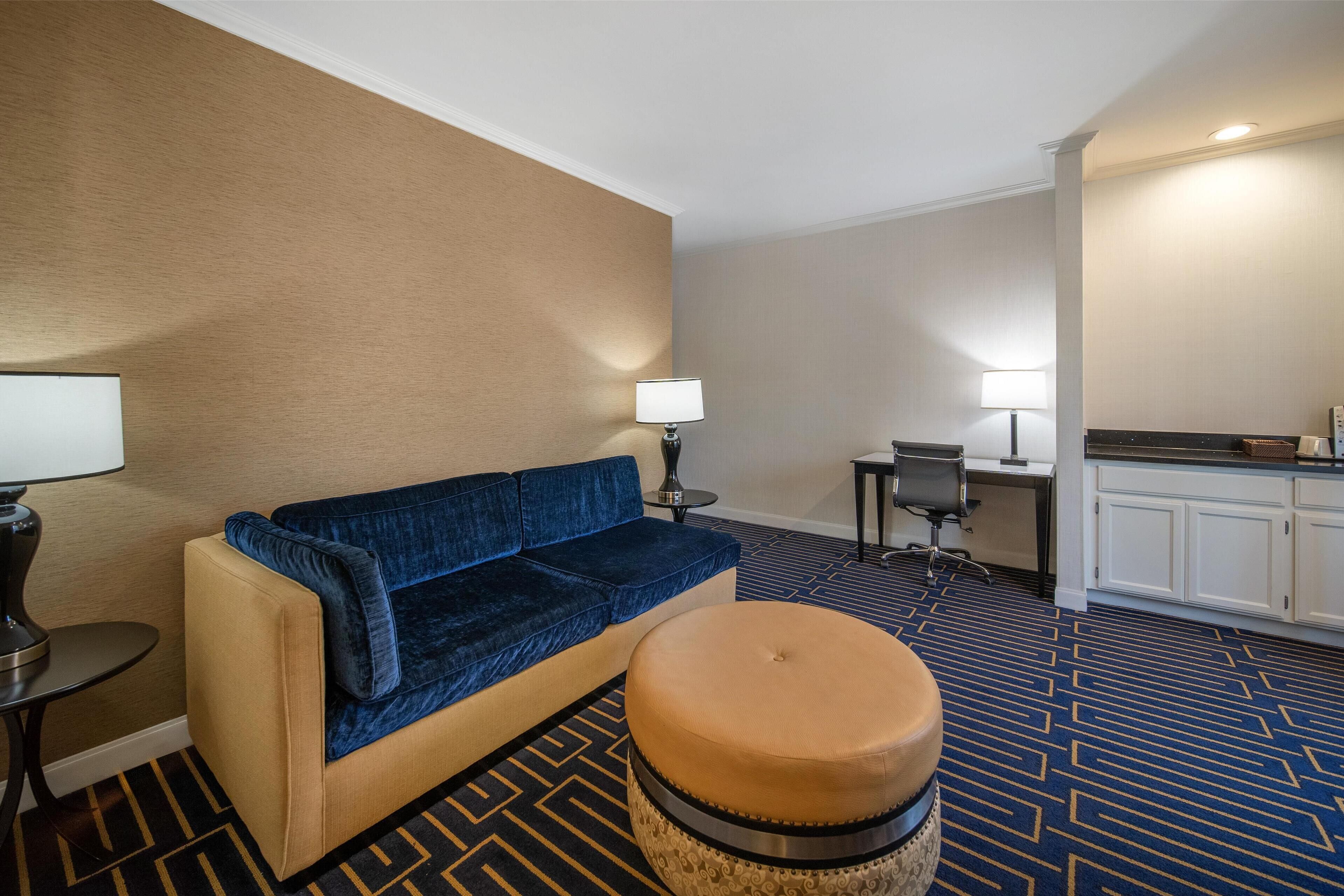 A hotel room with a sofa, ottoman, desk, chair, and a kitchenette area.