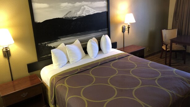 A hotel room with a large bed, a nightstand, a desk, and a chair. There is a painting on the wall and two lamps on either side of the bed. - Super 8 by Wyndham Corning (Corning)